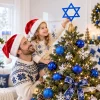 Father lifting his daughter to reach the blue Star of David topper on a tree decorated with blue balls and white snowflakes