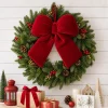13 inch red velvet bow centered at the top of an evergreen wreath with berries and pinecones on a white shiplap wall